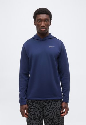 Joven con una sudadera con capucha Nike azul marino y pantalones negros con estampado de leopardo, posando frente a un fondo blanco liso.