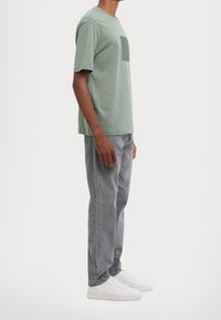 Světle zelené bavlněné tričko se čtvercovou náprsní kapsou, spárované se šedými rovnými džínami a bílými teniskami. Jednoduchý design a měkká textura.