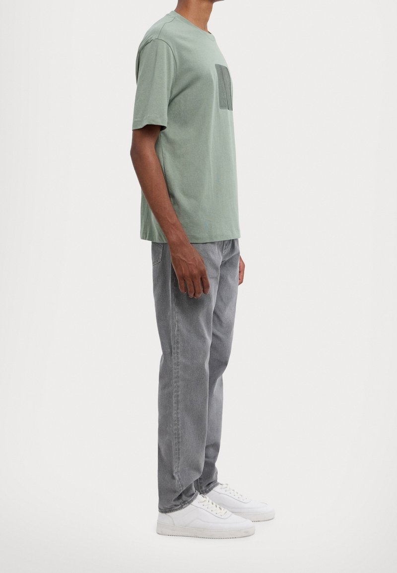 Světle zelené bavlněné tričko se čtvercovou náprsní kapsou, spárované se šedými rovnými džínami a bílými teniskami. Jednoduchý design a měkká textura.