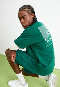 Grøn bomulds T-shirt med korte ærmer og et logo på ryggen; parret med matchende grønne shorts og hvide sneakers.