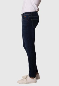 Jeans oscuros de corte slim con costuras visibles, bolsillos laterales y parche de piel en la parte trasera. Combinados con zapatillas blancas.