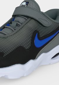 Κοντινό πλάνο σε γκρι αθλητικό παπούτσι Nike Air με μπλε λογότυπο, μαύρες λεπτομέρειες, πλέγμα ύφασμα, λευκή σόλα και γκρι λουράκι με βέλκρο πάνω από τα κορδόνια.