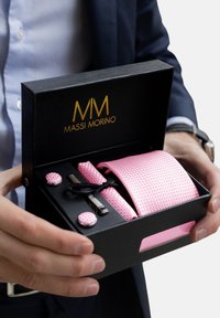 Rožinės tekstūruotos kaklaraištis, rankogalių mygtukai ir kaklaraiščio segtukas pateikti juodame dėkle su auksiniu ženklinimu. Kaklaraištis elegantiškai surištas su juostele.