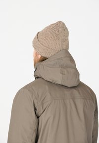 Bruine parka met een capuchon, met een gladde textuur en een losse pasvorm. G gedragen met een lichtbruine geribbelde breimuts.