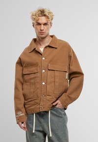 Braune Baumwolljacke mit Kragen, zwei Vordertaschen, silbernen Beschlägen und einer lockeren Passform. Getragen über hellgrauen Hose mit Kordelzug.