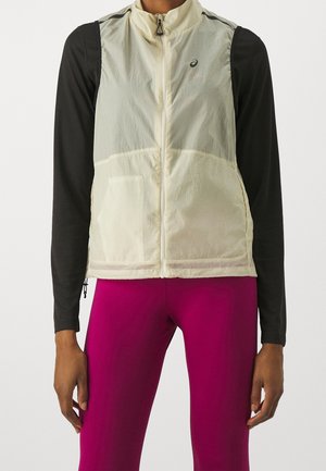 Personne portant un gilet beige sans manches à fermeture éclair sur une chemise noire à manches longues et un pantalon ajusté rose vif, debout les bras détendus.