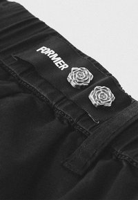 Le pantalon en denim noir présente une ceinture avec l'étiquette "FORMER", accentuée par deux ornements en forme de rose argentée de chaque côté.