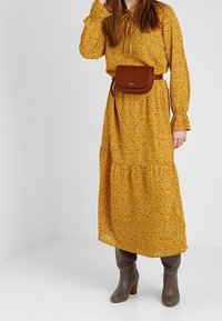Largo vestido floral amarillo con mangas largas y escote con lazo. Bolso cruzado marrón y botas altas marrones con tacón.