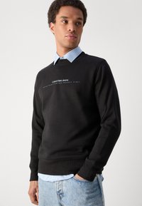 Crni pamučni dukserica s okruglim izrezom, rebrastim rukavima i strukom, s bijelim natpisom "Calvin Klein Jeans" ispod "Established Nineteen Seventy Eight."