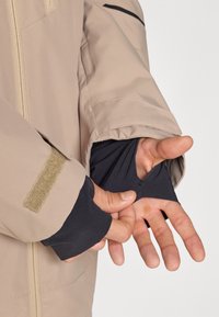 Beige szigetelt dzseki fekete belső bélésben, tépőzárral, cipzáros elülső résszel, és hüvelykujjnak nyílással ellátott ujjal.