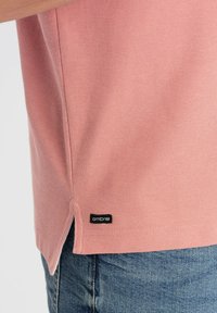 Světle růžové polo tričko s texturovanou látkou, bočními rozparky a černou etiketu s nápisem "ombre" na lemu. Džínové kraťasy viditelné na spodní části.