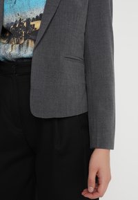 Blazer grigio con tessuto texture, chiusura a un bottone, rever a forma di notched e vestibilità slim. Abbinato a pantaloni scuri e una camicia a fantasia.