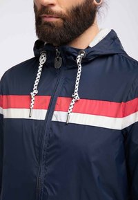 Námořnická větrovka s červenými a bílými vodorovnými pruhy, s předním zipem, kapucí na stažení a kontrastními kostičkovanými šňůrkami.