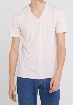 Mężczyzna ubrany w jasnoróżowy T-shirt z krótkim rękawem i dekoltem w serek oraz niebieskie dżinsy, stojący na jednolitym jasnym tle.