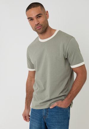 Mand med kort hår iført en salviegrøn T-shirt med hvide kanter, hænder i lommerne, stående mod en ensartet lys baggrund.