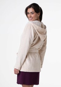 Robe con cappuccio color beige chiaro in tessuto texturizzato, con vita cinte e tasche frontali, abbinata a una gonna plissettata viola. Design semplice e funzionale.