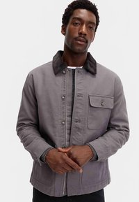 Grijze jas met een kraag van ribfluweel, knoop- en ritssluiting, voorzak en geribbelde manchetten. De stof oogt duurzaam en gestructureerd.