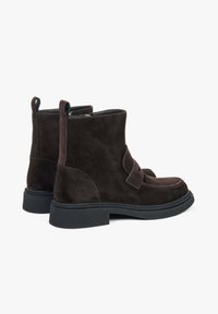 Pár tmavě hnědých semišových kotníkových bot s černými výraznými podrážkami a poutky na zadní straně, zobrazené ze zadního úhlu na bílém pozadí.