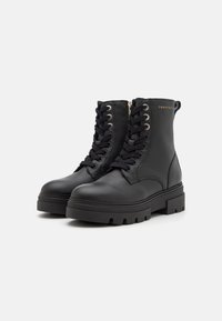Une paire de bottes noires en cuir à la cheville avec des semelles épaisses, un laçage à l'avant, des zips latéraux et un logo de marque doré discret sur le côté extérieur.
