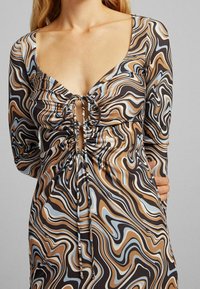 Žena v šatech s dlouhým rukávem, které mají hnědé, černé, béžové a modré vlnité vzory a řasený přední díl stažený šňůrkami.