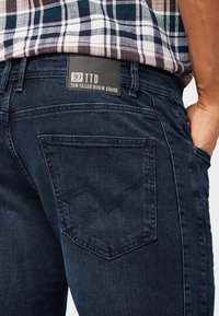 Vista trasera de una persona que lleva jeans de mezclilla azul oscuro con la etiqueta de la marca Tom Tailor Denim y una camisa de cuadros en azul, blanco y rojo.