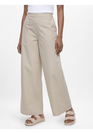 Beige broek met wijde pijpen gecombineerd met lichtbeige sandalen met dubbele gesp, gedragen door een persoon die staat tegen een effen witte achtergrond.