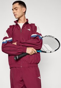 Bărbat în trening bordo ținând o rachetă de tenis încrucișată pe piept, uitându-se într-o parte, pe un fundal simplu.