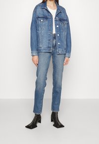 Chaqueta de denim azul claro con dos bolsillos en el pecho y herrajes plateados. Combinada con jeans azules y botas de tacón negro hasta el tobillo.