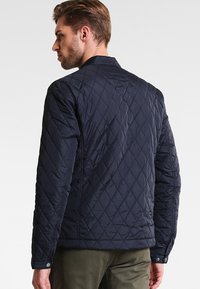 Mörkblå vadderad jacka med ståkrage, som har diamantmönstrad sömnad och tryckknappar vid ärmsluten. Texturerat nylonmaterial.