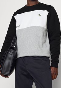 Persona che indossa un felpa Lacoste nera, bianca e grigia, tiene una borsa a spalla in pelle nera, vestita con pantaloni scuri.