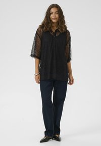 Blouse en dentelle noire avec un col et des manches mi-longues, associée à un jean bleu foncé et des chaussures plates noires. Coupe ample et texturée.