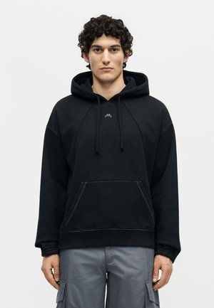 Mladić s kovrčavom kosom nosi crnu majicu s kapuljačom i sive cargo hlače, stoji na bijeloj pozadini.