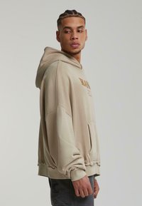 Haut à capuche beige avec un cordon de serrage, coupe oversize, tissu texturé et poche avant. Présente un logo imprimé dans un beige plus foncé.