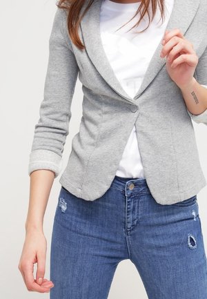 Femme portant un blazer gris clair cintré sur une chemise blanche et un jean bleu déchiré, avec les manches retroussées et un tatouage visible sur le bras.