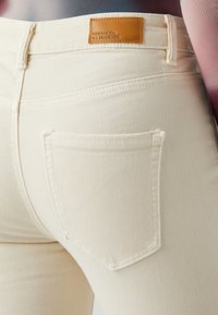Jeans de denim de culoare crem au o croială structurată, cu două buzunare la spate și o etichetă din piele la talie. Textura pare netedă.