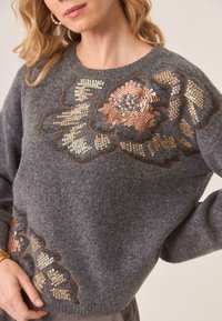 Suéter de lana gris con bordado de lentejuelas florales en tonos dorados y rosas, cuello redondo y corte holgado. Patrón de punto texturizado.