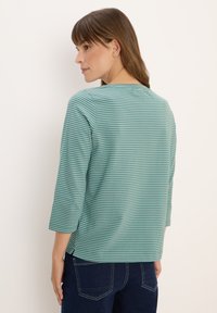 Femme aux cheveux bruns portant un haut à manches trois-quarts rayé vert et blanc et un jean bleu foncé, vue de profil contre un fond uni.