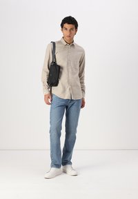 Cămașă bej cu nasturi, blugi albaștri deschiși și adidași albi. Geantă neagră tip crossbody. Design simplu, croi lejer, textură de țesătură netedă.