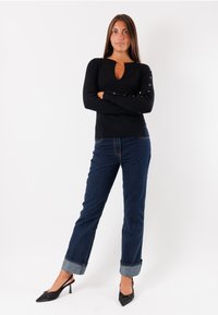 Top nero a coste con maniche lunghe, scollatura a chiave e dettagli in bottone argentato sulle maniche, abbinato a jeans scuri in denim con risvolti e tacchi neri.