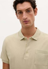 Polo de hombre en color beige claro hecho de suave algodón. Presenta un cuello, una tapeta de tres botones y un pequeño bolsillo en el pecho con acento acanalado.