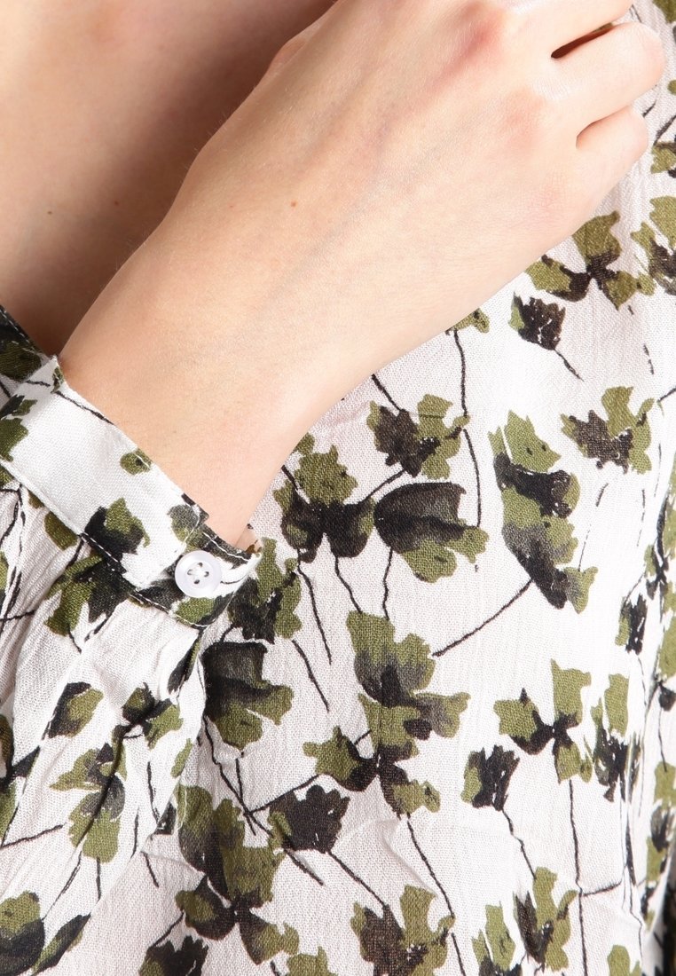 Gros plan sur un poignet et une main portant un chemisier blanc à imprimé floral vert et noir avec un poignet blanc boutonné.