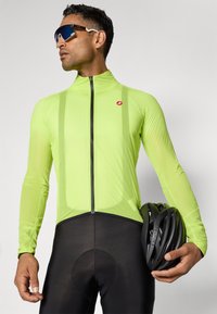 Neonowy zielony rowerowy jacket z długim rękawem i czarnym zamkiem, w połączeniu z czarnymi szortami rowerowymi z wkładką amortyzującą; kask trzymany w ręku.