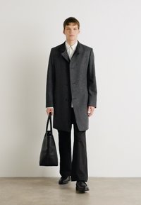 Grau Wollmantel mit Reverskragen, drei Knöpfen und seitlichen Taschen. Wird mit schwarzen Hosen getragen und hält eine schwarze Leder-Tote.