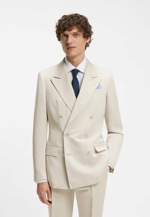 Άνδρας με σγουρά μαλλιά που φοράει εκρού σακάκι διπλής σειράς κουμπιών, λευκό πουκάμισο, ναυτικό μπλε γραβάτα και μαντηλάκι τσέπης με σχέδιο, ποζάροντας μπροστά σε λευκό φόντο.