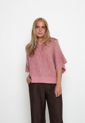Vrouw met lang blond haar die een losse roze zachte trui en donkere wijde broek draagt, staand met één hand in de zak.