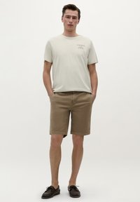 Tricou beige deschis cu mâneci scurte și imprimeu text, purtat împreună cu o pereche de pantaloni scurți maro. Pantalonii scurți au o croială clasică și o textură netedă. Încălțăminte închisă completează aspectul.