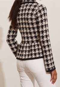 Mustavalkoinen kuviollinen neuletakki, jossa on peplum-vyötärö, pitkät hihat ja kaulus, yhdistetty valkoisiin housuihin. Teksturoitu kangasosa näkyvissä.