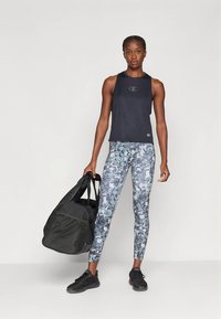 Svart ermeløs treningssinglet med logo, kombinert med leggings med kamuflasjemønster i nyanser av grått og blått. Modellen holder en svart duffelbag.