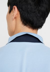 Polo bleu clair avec un col bleu marine, présentant un tissu texturé et des accents contrastés au niveau de l'encolure.