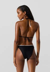 Set di bikini neri con top a collarino con chiusura a fiocco e parte inferiore a vita alta con dettagli a scallop bianchi. Texture liscia.